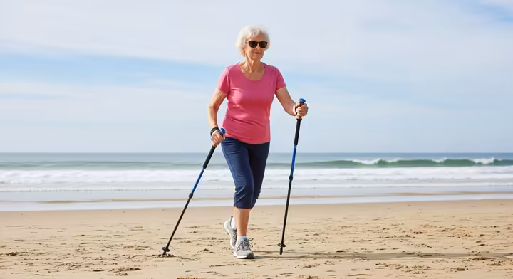 A senior confidently walking on a sandy beach using a pair of walking poles for extra stability.